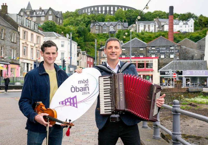 Two men, one holding a Mod Oban sign and the other holding an Accordion, on Oban seafront.